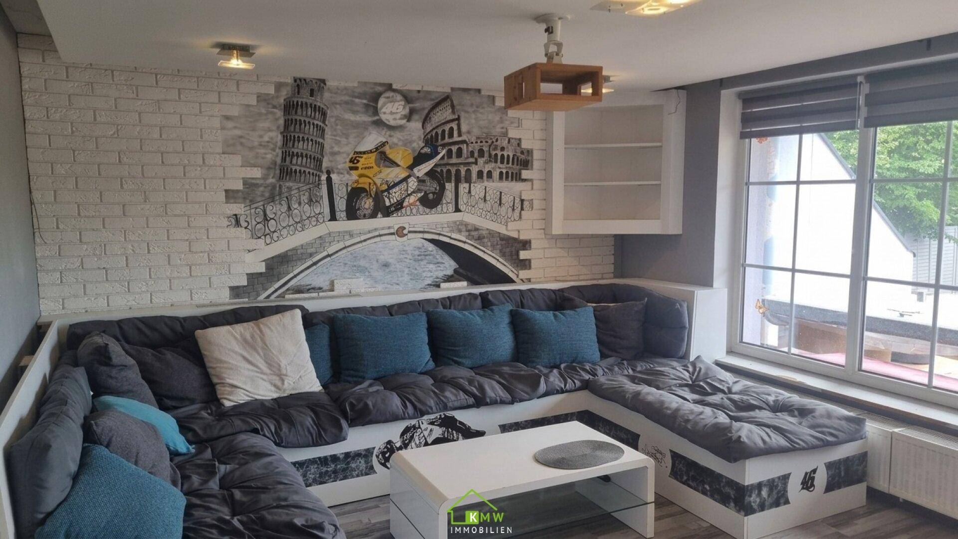 Gemütliches Wohnzimmer mit großer Eckcouch, Wandgemälde und einem Fenster mit Blick ins Grüne.