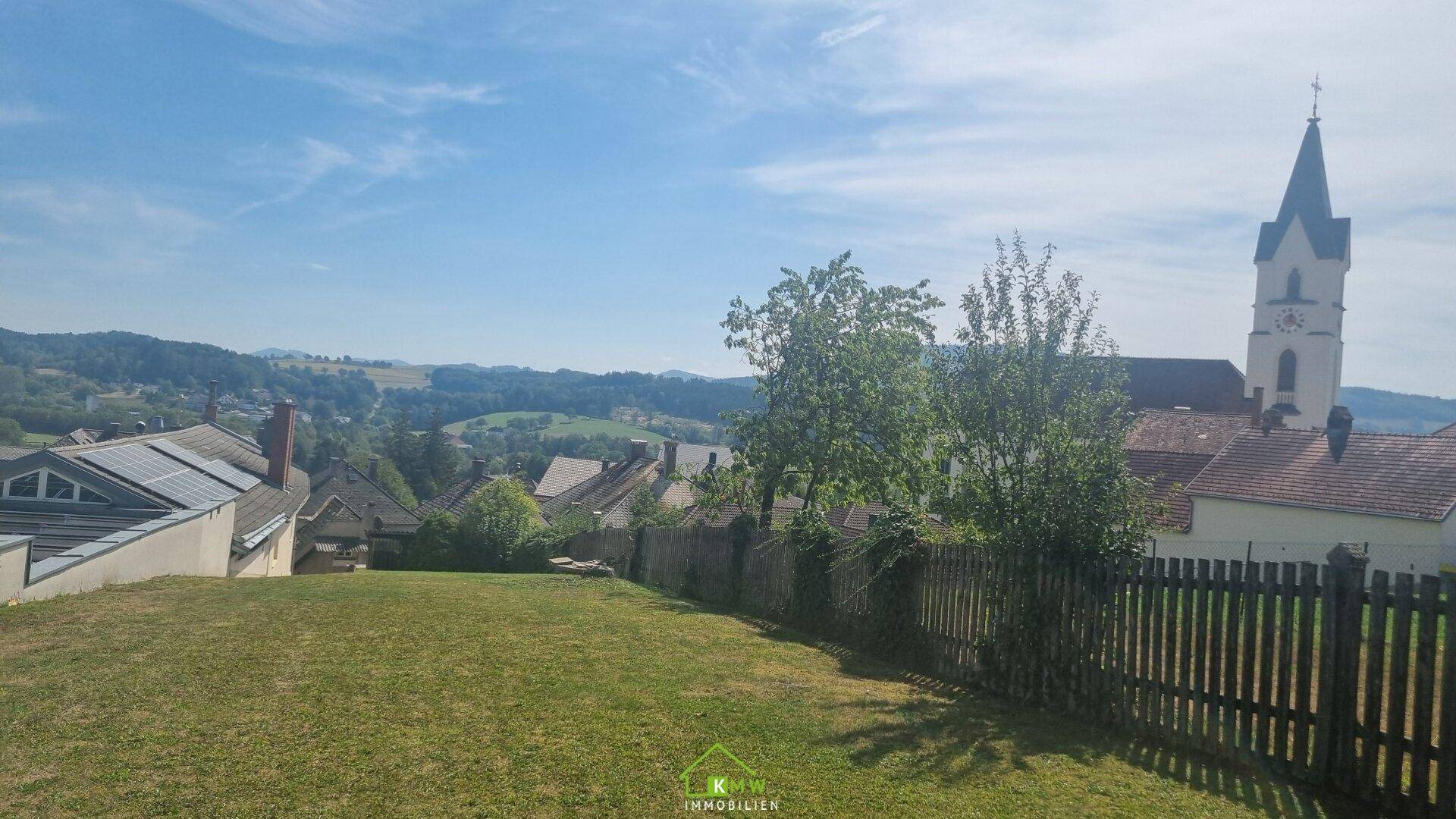 Weitläufiger Blick von einer Rasenfläche auf ein Dorf mit Kirchturm und sanfte Hügel in der Ferne.