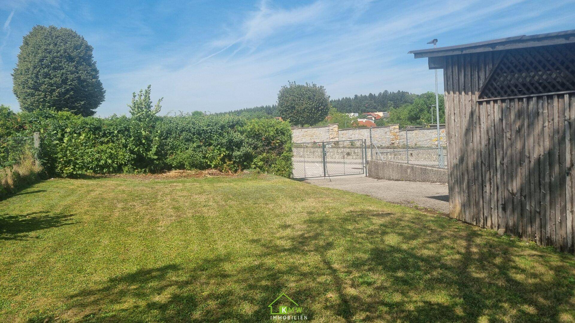 Grüne Rasenfläche mit einer Hecke, einem Holzschuppen und einem Zaun, umgeben von Natur.