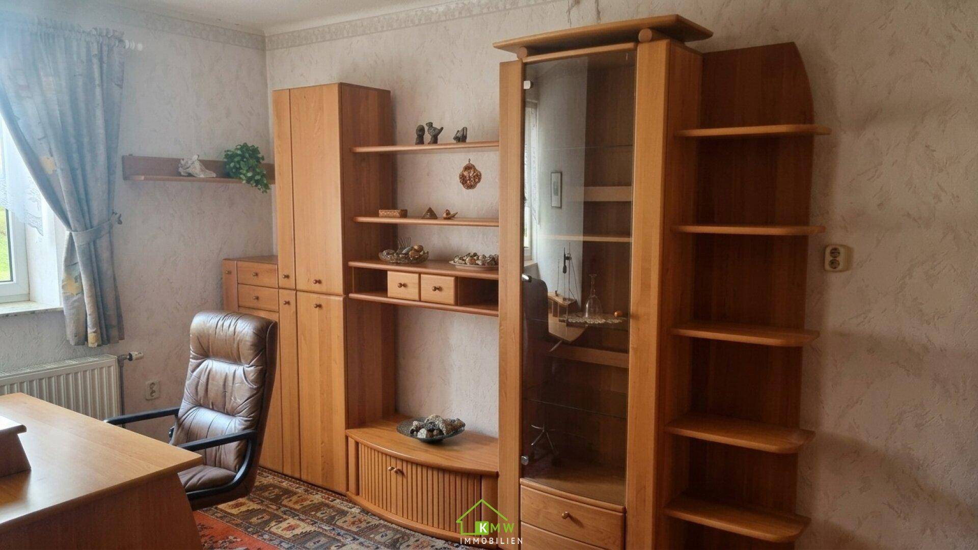 Wohnzimmer mit Holzmöbeln, einem Schreibtisch und einem bequemen Sessel, dekoriert mit gemusterter Tapete.