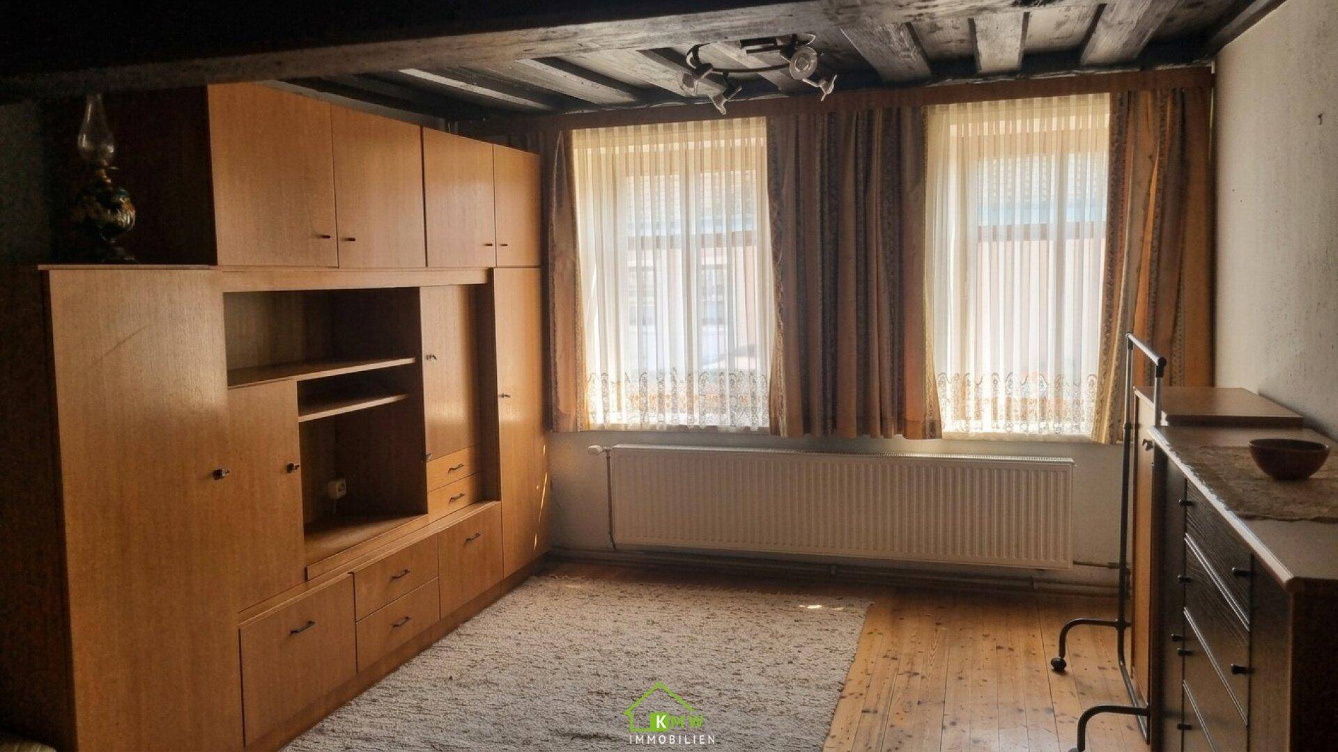 Zimmer mit großen Holzschränken, zwei Fenstern und einem Teppich auf Holzdielenboden, mit sichtbaren Deckenbalken.