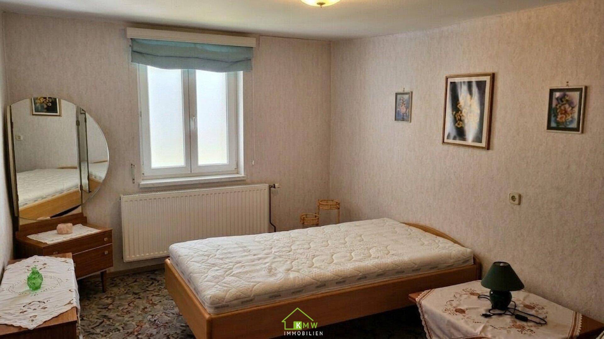 Schlafzimmer mit Einzelbett, Fenster und einem Schminktisch mit Spiegel, dekoriert mit gemusterter Tapete.
