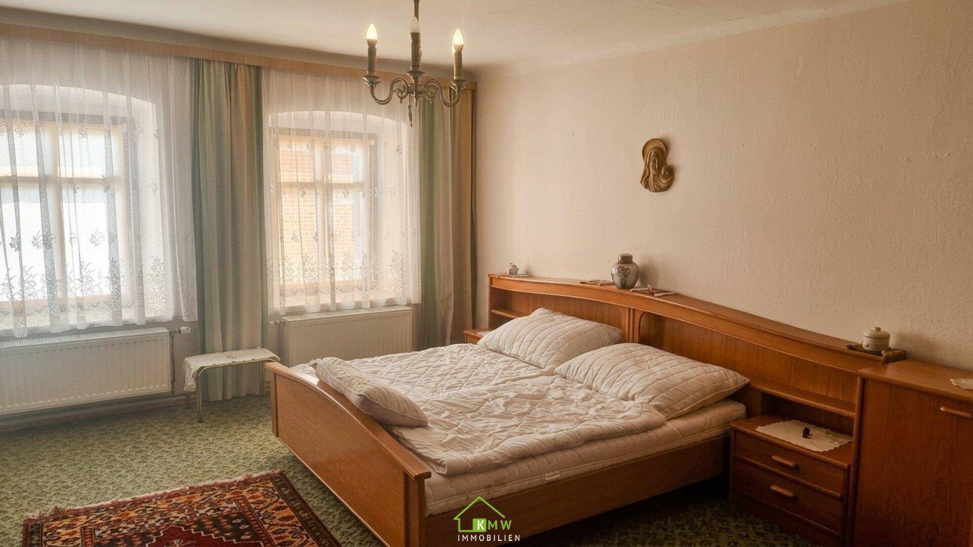 Geräumiges Schlafzimmer mit Doppelbett, zwei Fenstern und einem Kronleuchter, eingerichtet im klassischen Stil.