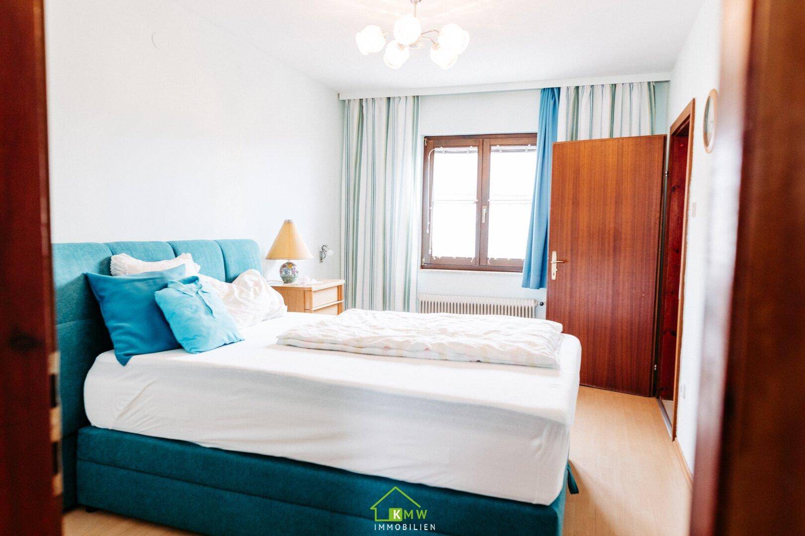 Gemütliches Schlafzimmer mit blauem Polsterbett, hellem Laminatboden und Fenster mit Vorhängen.