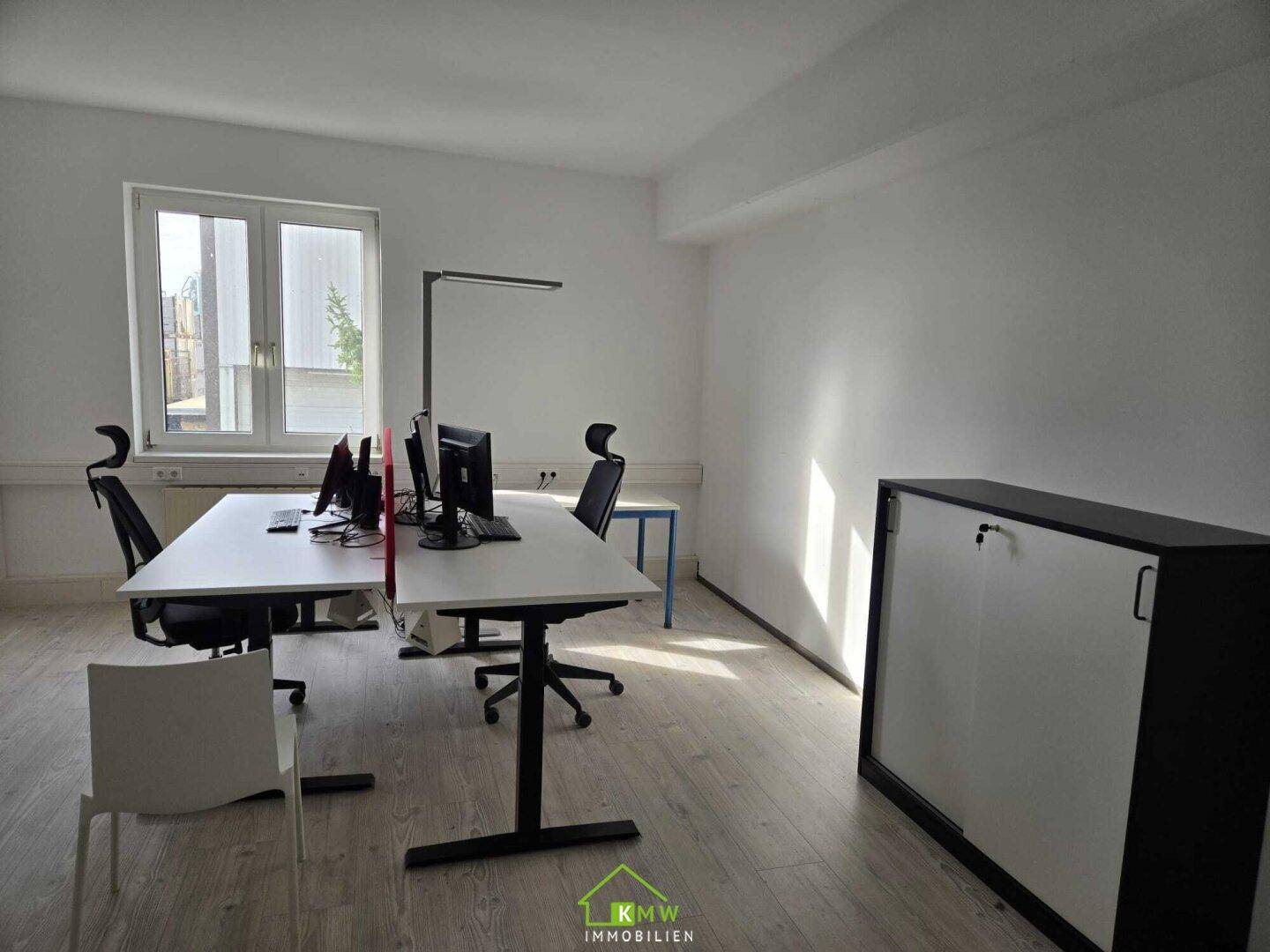 Geräumiges Büro mit mehreren Arbeitsplätzen, großen Fenstern und hellem Laminatboden.