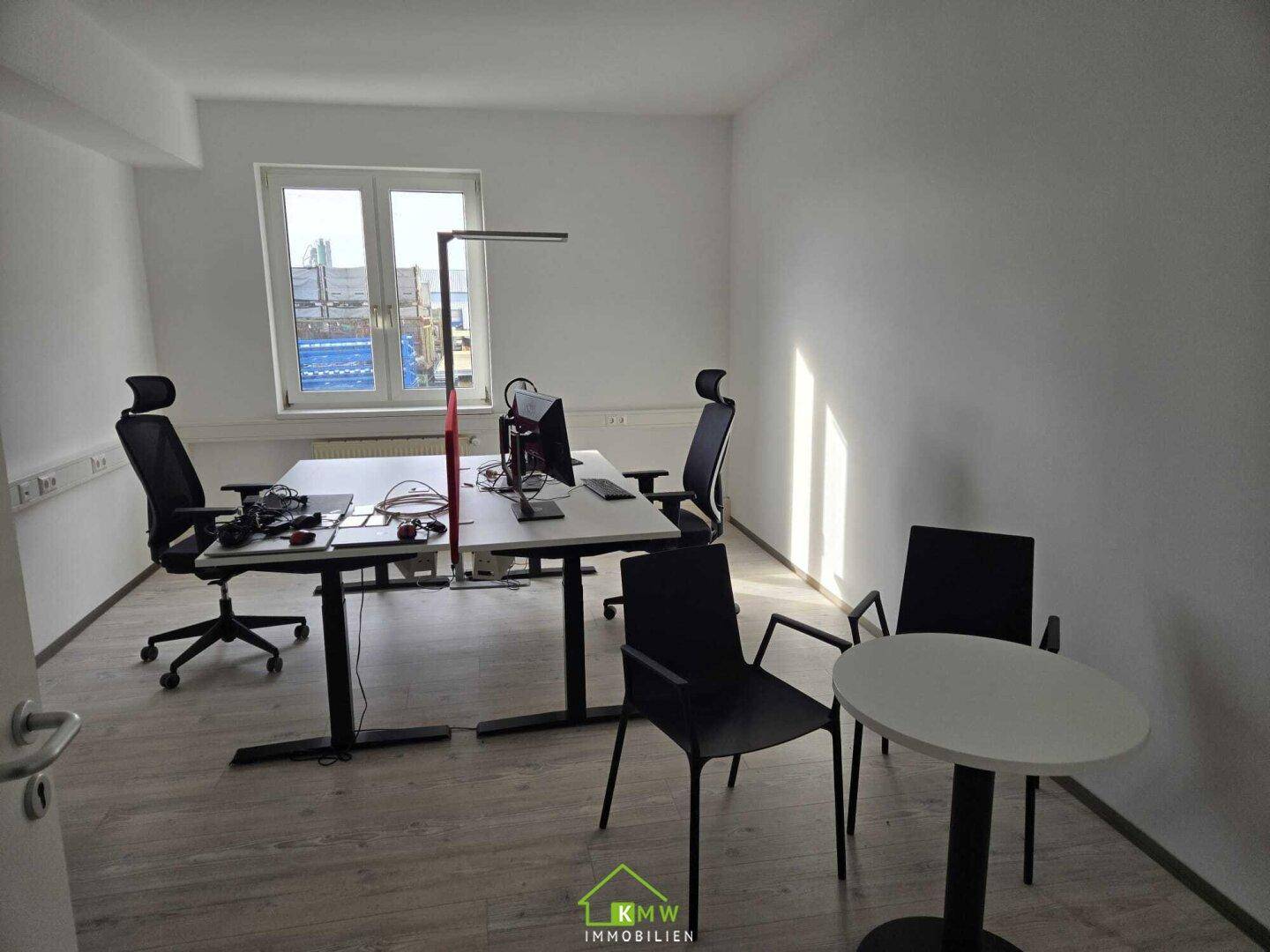 Modernes Büro mit mehreren Arbeitsplätzen und einem kleinen Besprechungstisch am Fenster.