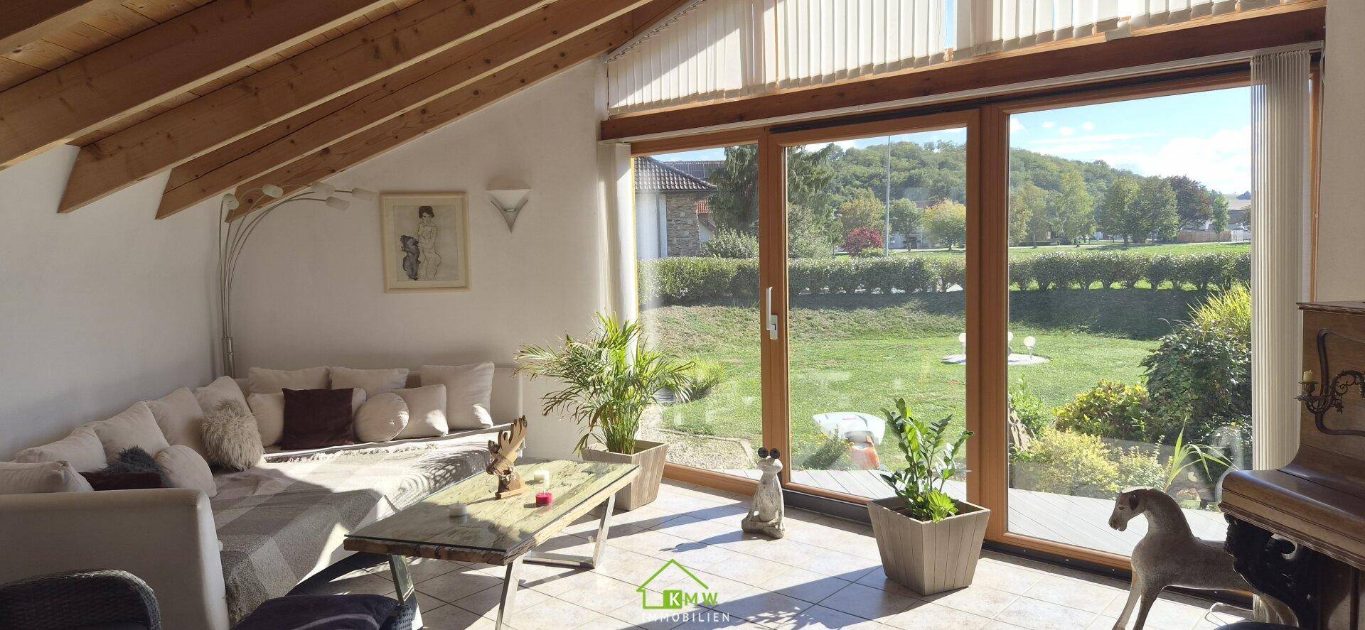 Lichtdurchflutetes Wohnzimmer mit großen Fenstern, Holzbalkendecke und Gartenblick.