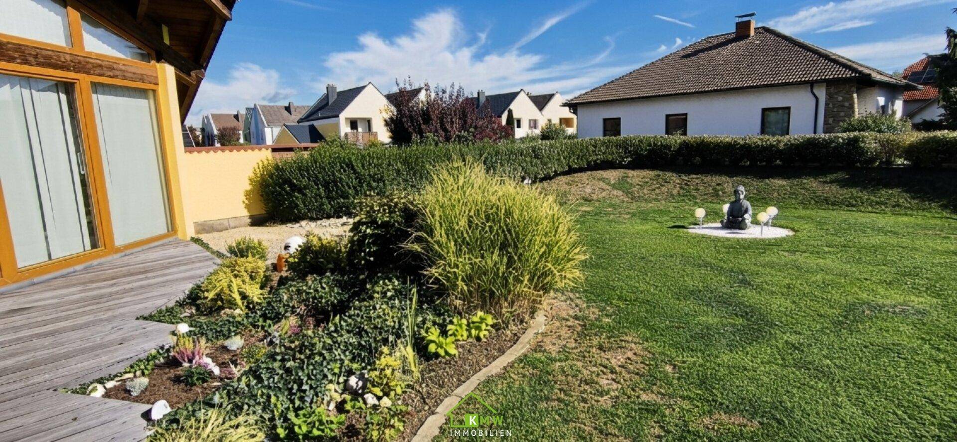 Gartenbereich mit Holzterrasse, Rasen und dekorativen Pflanzen unter blauem Himmel.