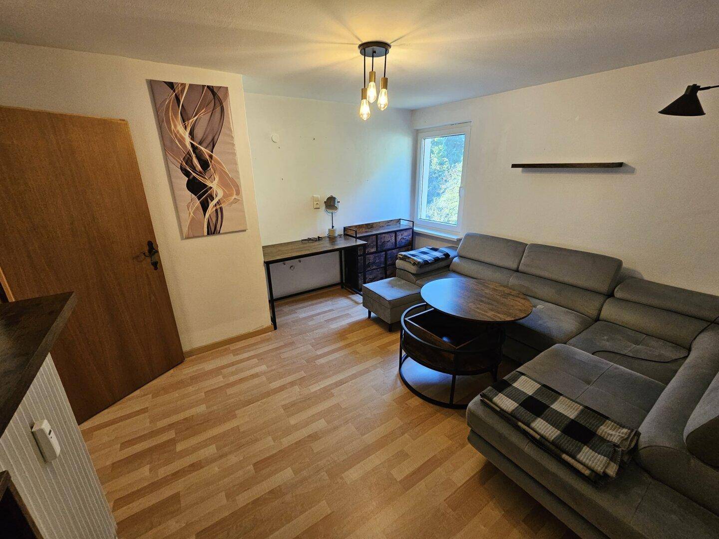 Gemütliches Wohnzimmer mit Ecksofa, Holzboden und Fenster mit Blick ins Grüne.