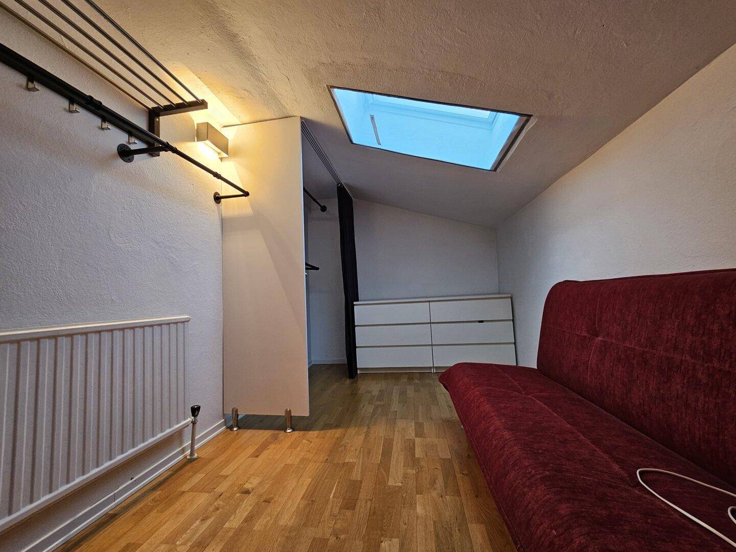 Raum mit rotem Sofa, Dachfenster und offener Kleiderstange unter schräger Decke.