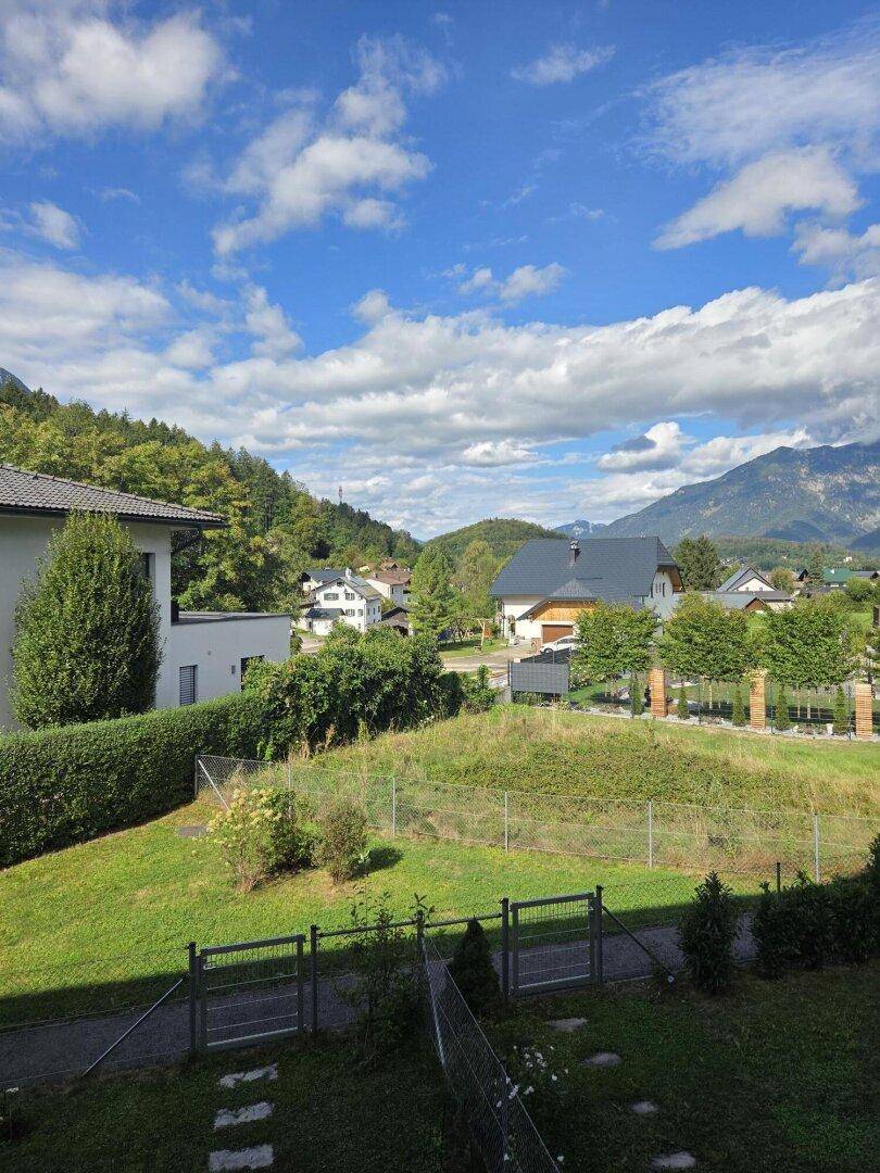 Weitläufiger Ausblick auf eine Wohnsiedlung, grüne Hügel und majestätische Berge unter blauem Himmel.