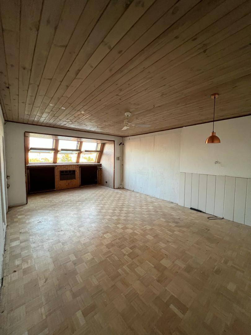 Geräumiges Wohnzimmer mit Parkettboden, Holzdecke und breiter Fensterfront, die viel Licht spendet.