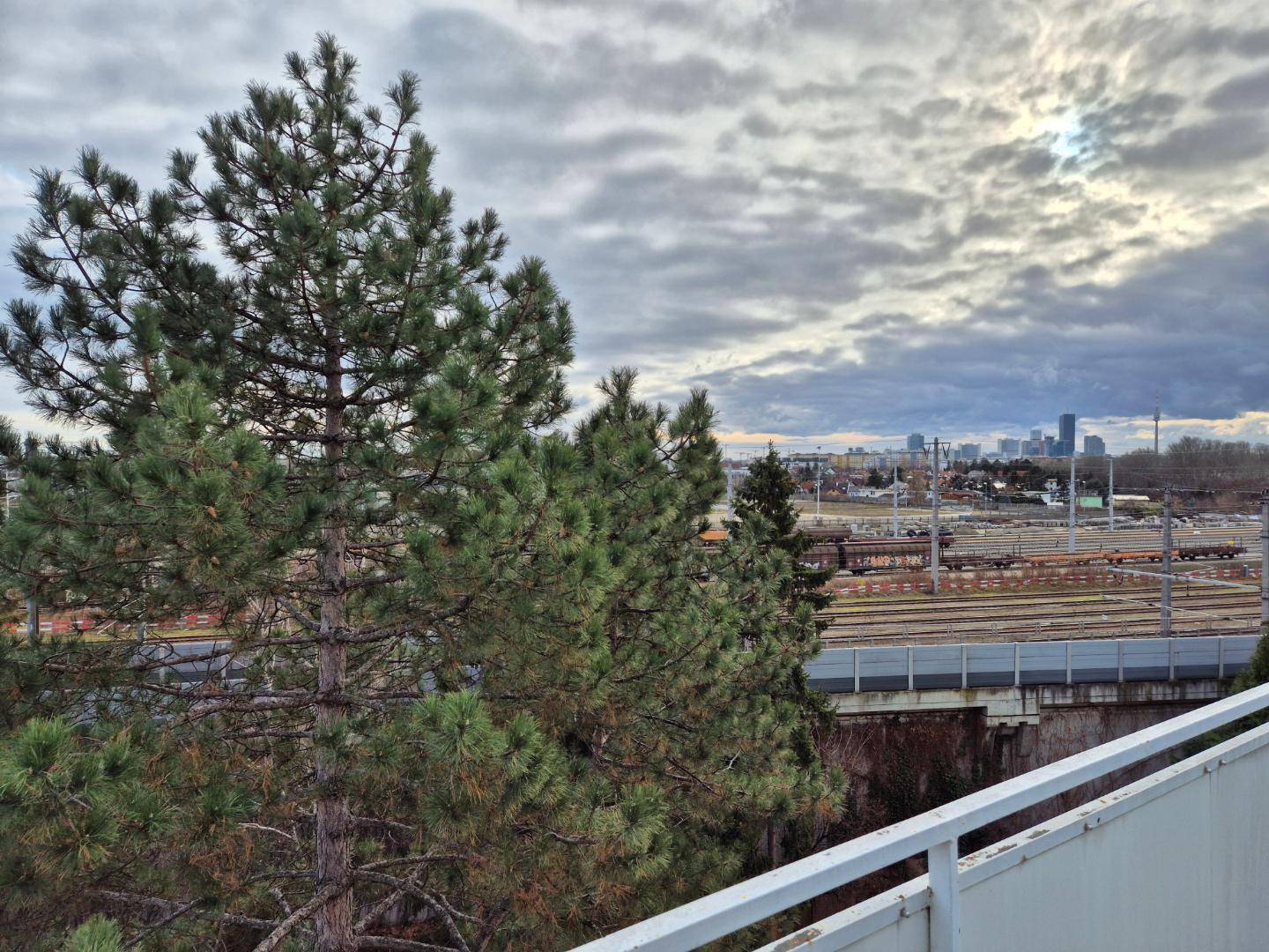 Ausblick vom Balkon auf Bahngleise, Bäume und die Skyline der Stadt am Horizont unter bewölktem Himmel.