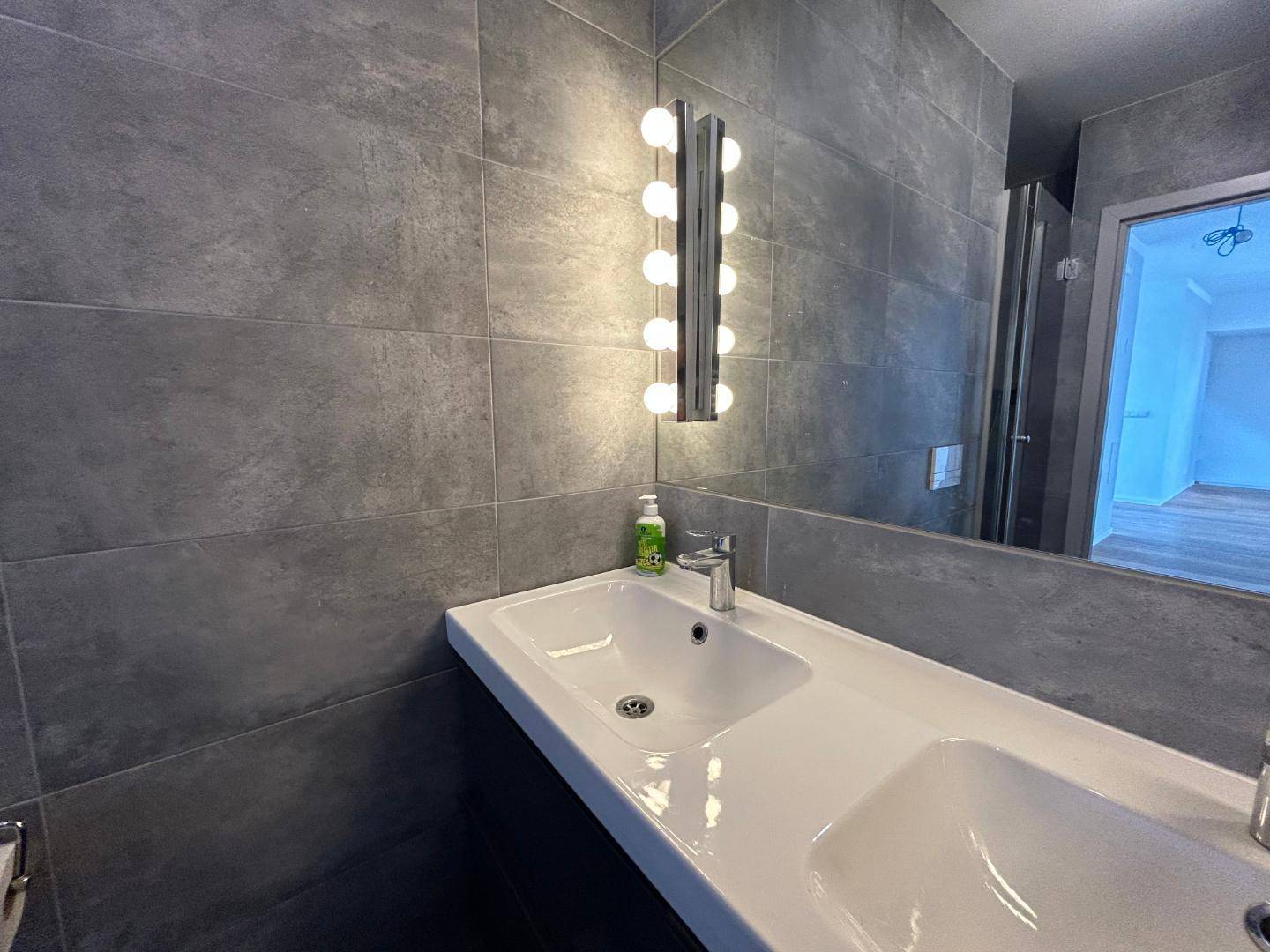 Modernes Badezimmer mit grauen Fliesen, Doppelwaschbecken und großem Spiegel mit Beleuchtung.