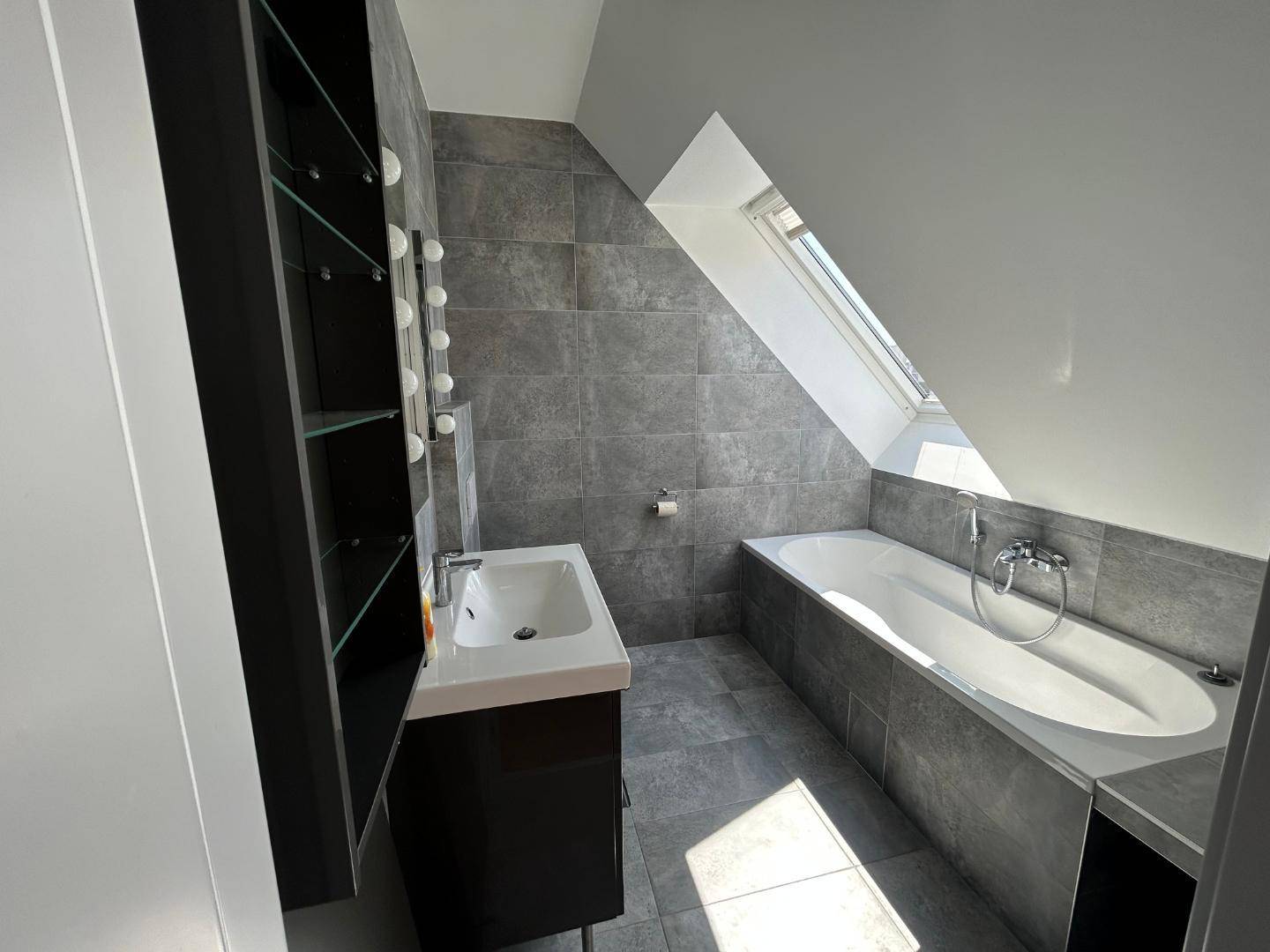 Modernes Badezimmer mit Badewanne unter Dachschräge, Dachfenster und grauen Fliesen.