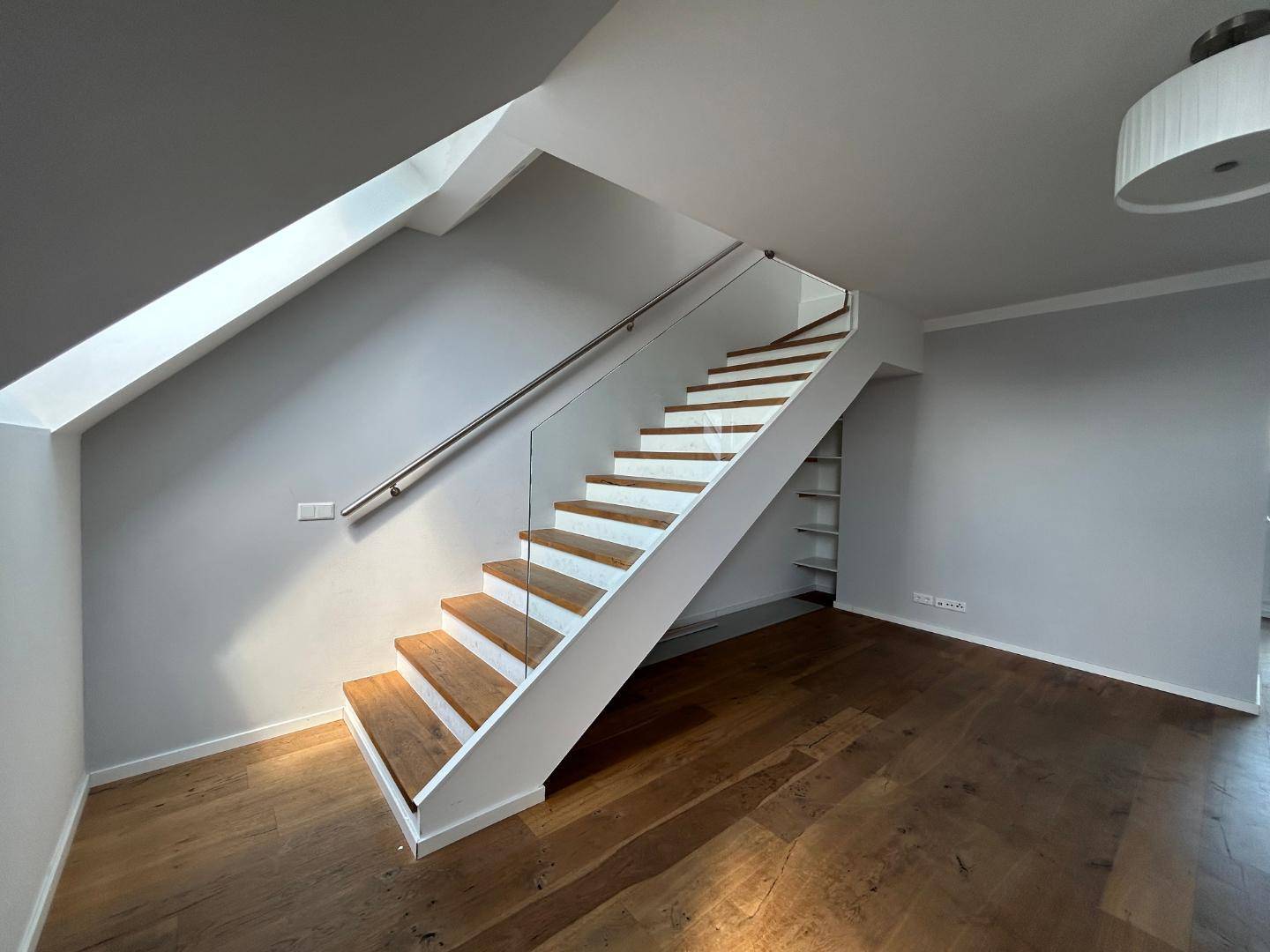 Moderne Holztreppe mit Glasgeländer und schrägen Wänden, die in das obere Stockwerk führt.