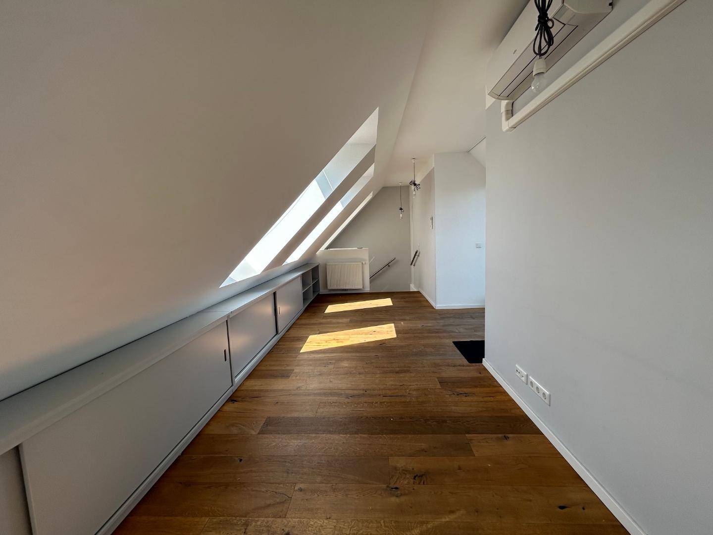 Geräumiger Raum mit Holzdielenboden, schrägen Decken, Dachfenstern, Stauschränken und Klimaanlage.