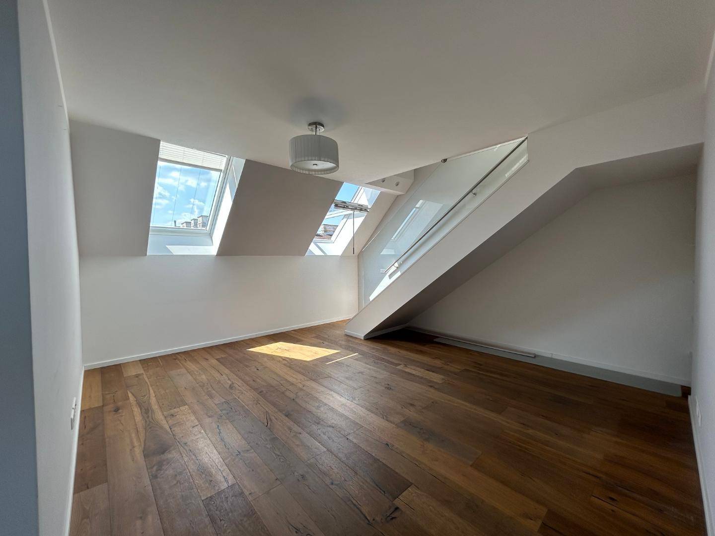 Heller Wohnbereich mit Holzdielenboden, schrägen Decken, mehreren Dachfenstern und moderner Treppe mit Glasgeländer.