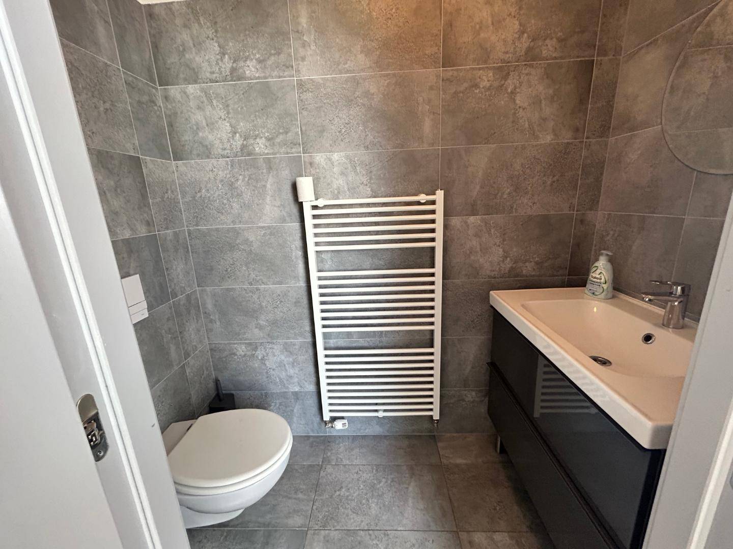 Modernes Gäste-WC mit grauen Fliesen, wandhängendem WC, Einzelwaschbecken und beheiztem Handtuchhalter.