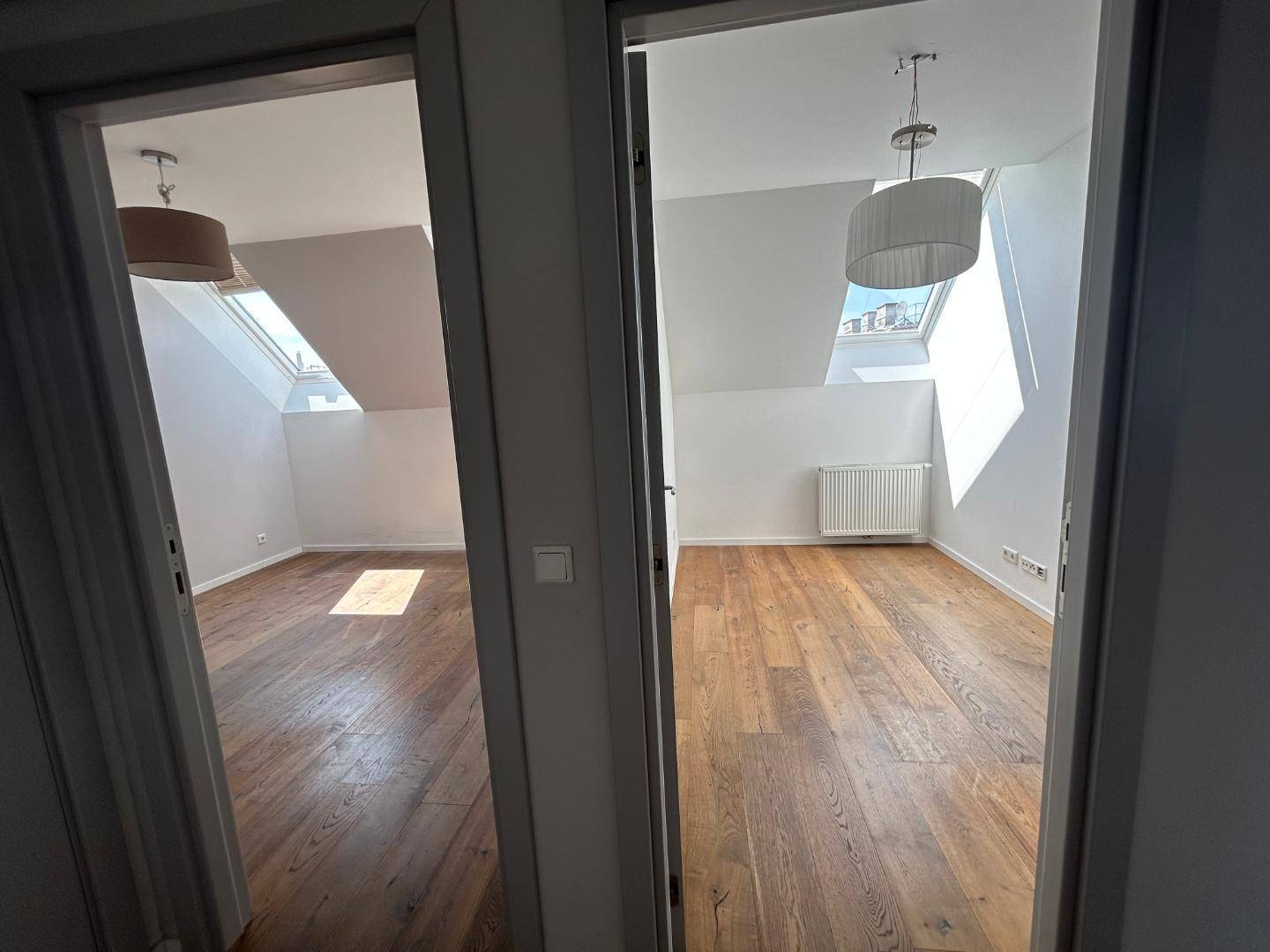 Blick in zwei helle Räume mit Holzdielenböden, schrägen Decken und großen Dachfenstern.