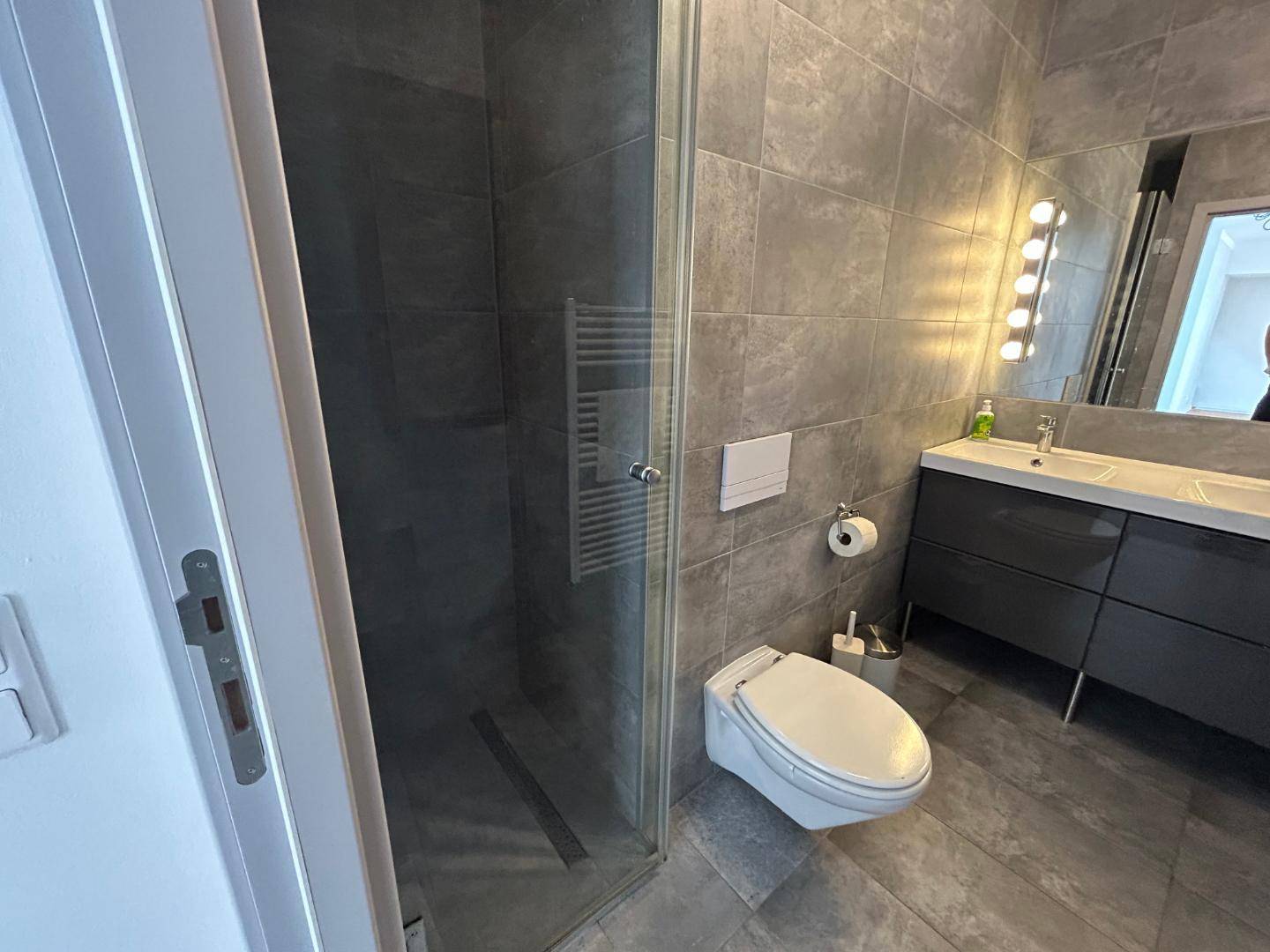 Modernes Badezimmer mit grauen Fliesen, wandhängendem WC, Doppelwaschbecken und begehbarer Dusche.