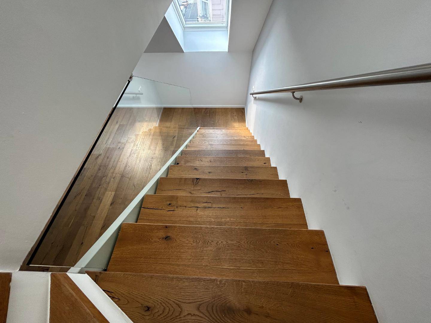 Blick nach unten auf die moderne Holztreppe mit Glasgeländer und einem Dachfenster im oberen Bereich.