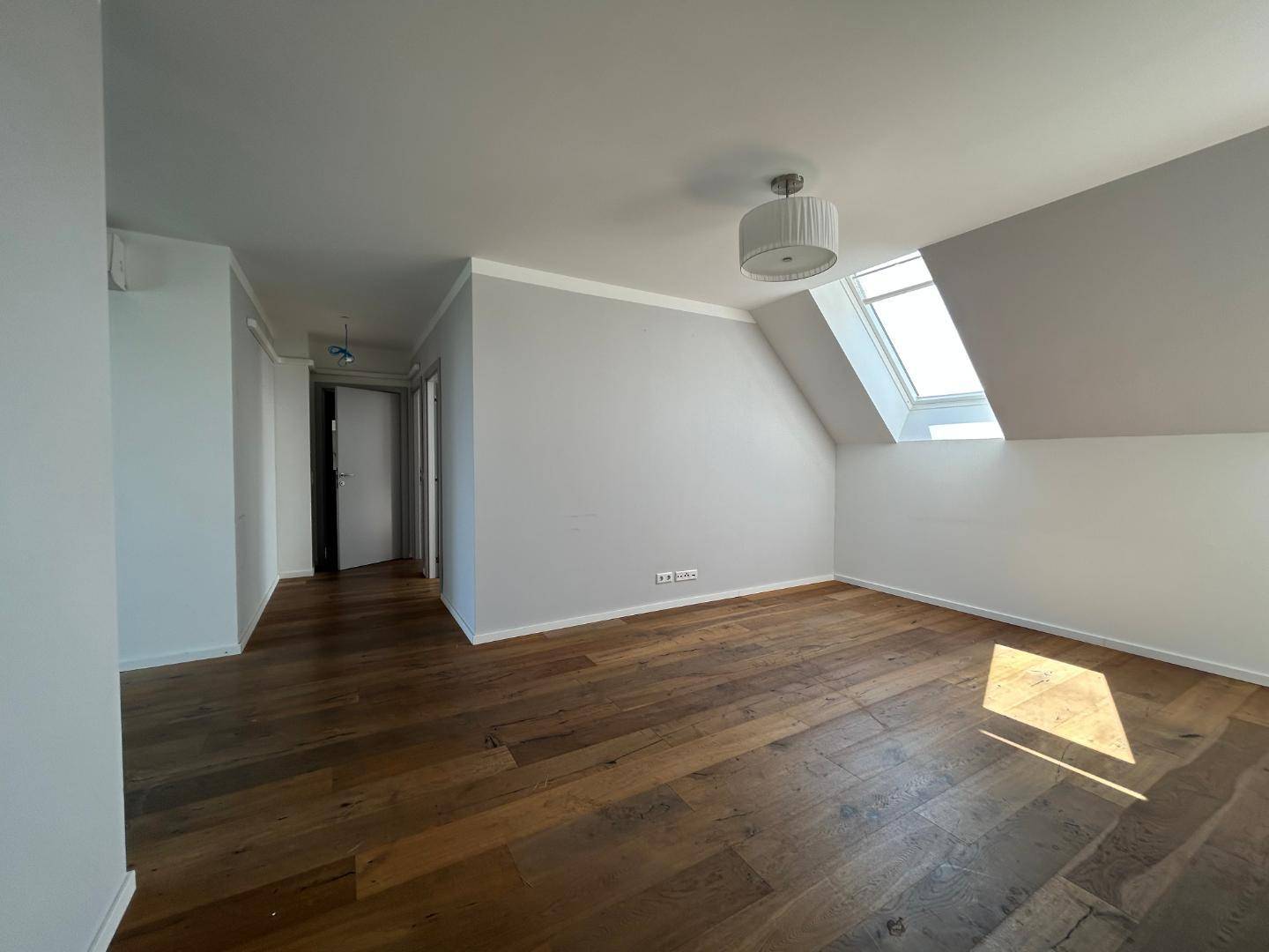 Geräumiger Wohnbereich mit Holzdielenboden, weißen Wänden, schräger Decke und einem großen Dachfenster.