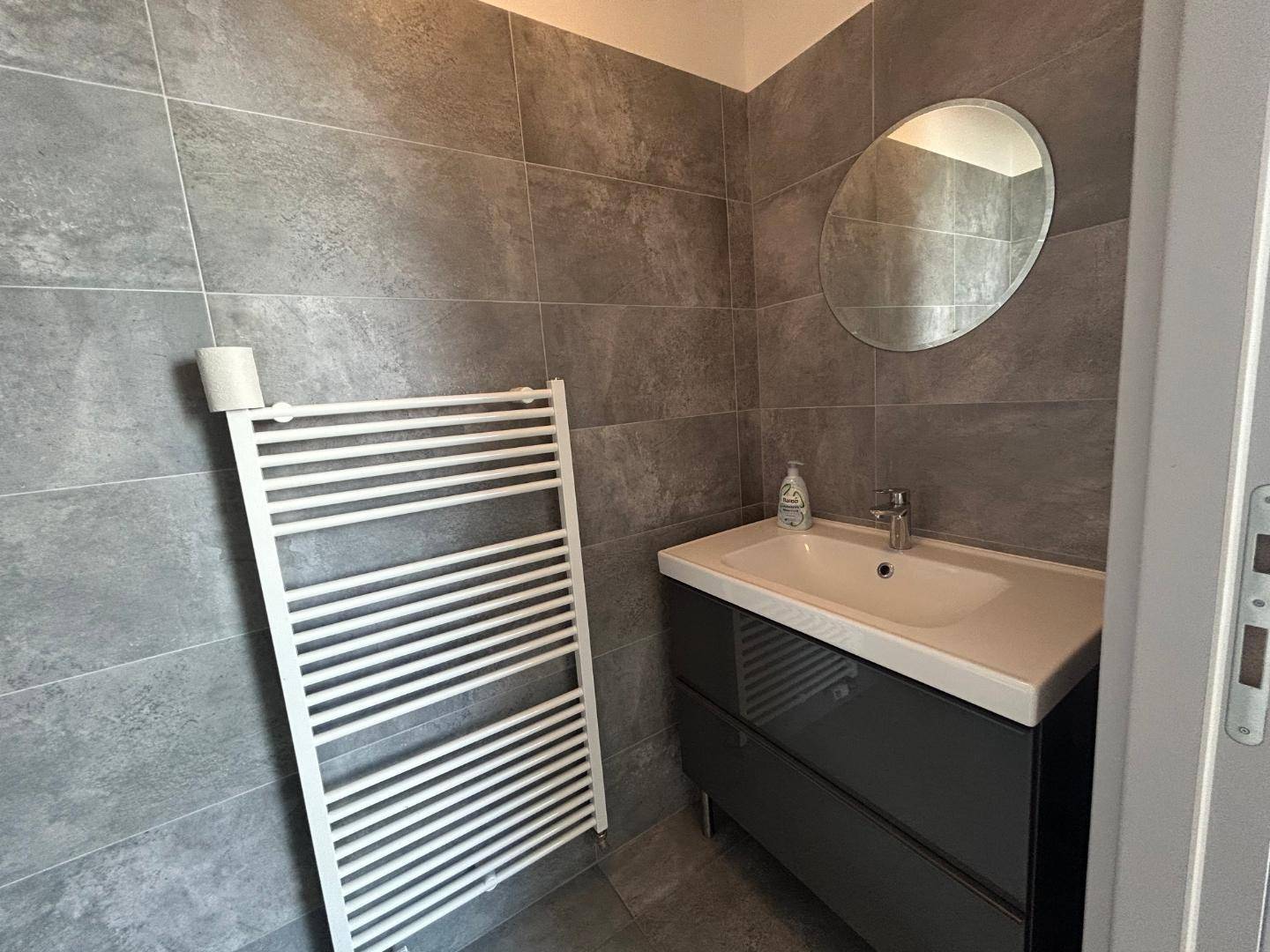 Modernes Gäste-WC mit grauen Fliesen, Einzelwaschbecken, rundem Spiegel und beheiztem Handtuchhalter.