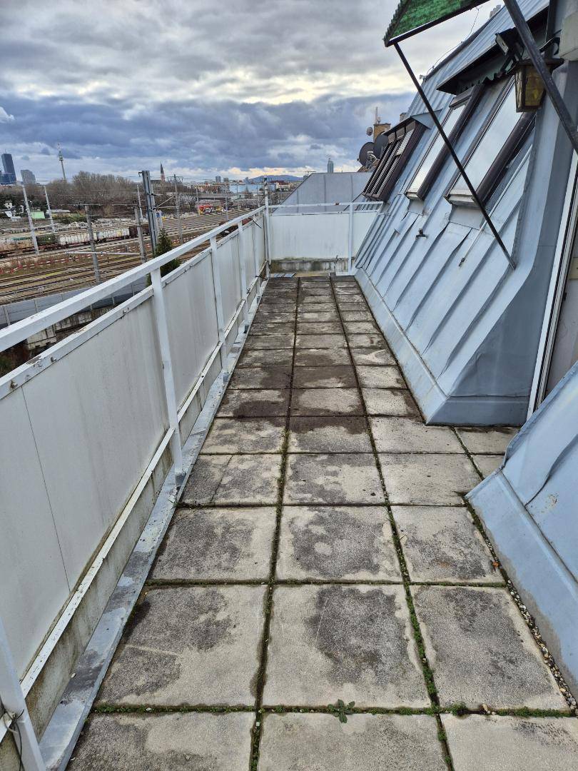 Langer, gefliester Balkon mit Metallgeländer und Blick auf die Stadt, in renovierungsbedürftigem Zustand.