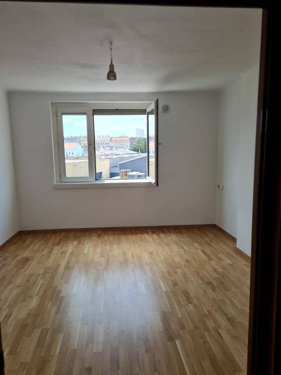 Heller, leerer Raum mit Holzparkettboden und einem geöffneten Fenster, das einen Blick nach draußen bietet.