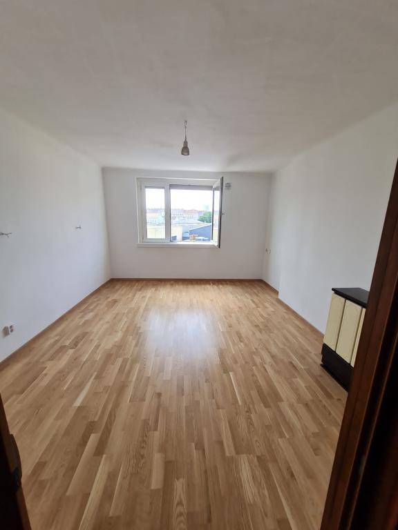 Leerer Raum mit hellem Holzparkettboden und einem Fenster, das Tageslicht hereinlässt.