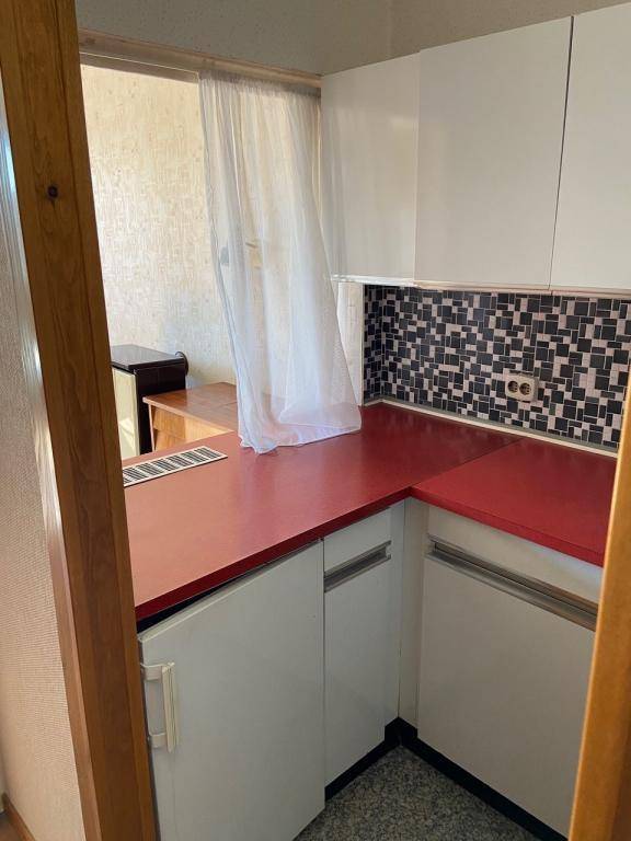 Kleine Küche mit roten Arbeitsplatten, weißen Schränken und einem Fenster mit Vorhang.