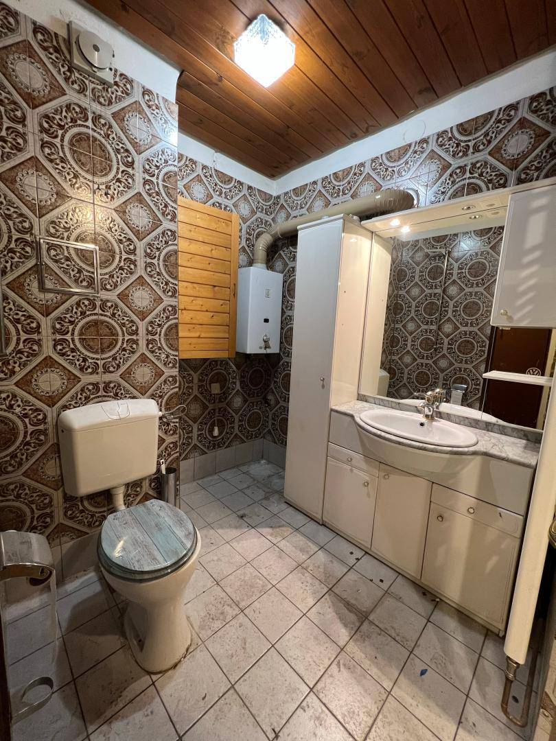 Veraltetes Badezimmer mit gemusterten Fliesen, Holzdecke, Toilette und einem einzelnen Waschbecken.