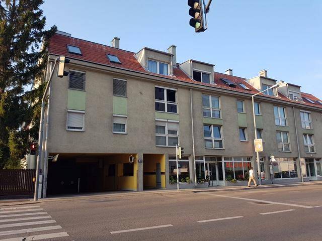 Mehrfamilienhaus mit rotem Dach, Dachgauben und Geschäften im Erdgeschoss an einer Straße.