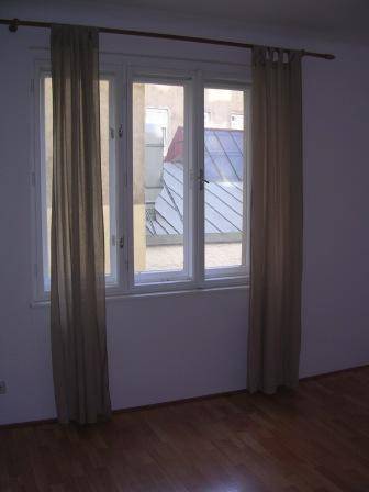 Fenster mit beigen Vorhängen und Blick auf ein gegenüberliegendes Dach.
