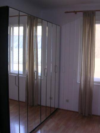 Schlafzimmer mit großem Spiegelschrank, Holzfußboden und Fenstern mit beigen Vorhängen.