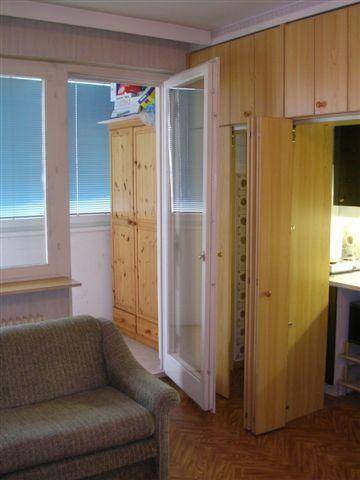 Wohnzimmer mit Sofa, Fenster mit Jalousien und Zugang zur versteckten Kochnische durch Falttüren.