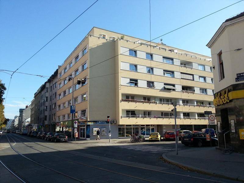 Mehrstöckiges Wohngebäude mit Balkonen und Geschäften im Erdgeschoss an einer Stadtstraße.