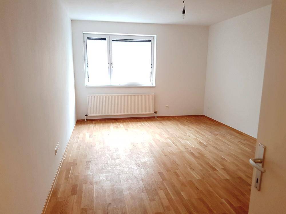 Helles, leeres Zimmer mit Parkettboden, weißen Wänden und einem Fenster mit Heizkörper.