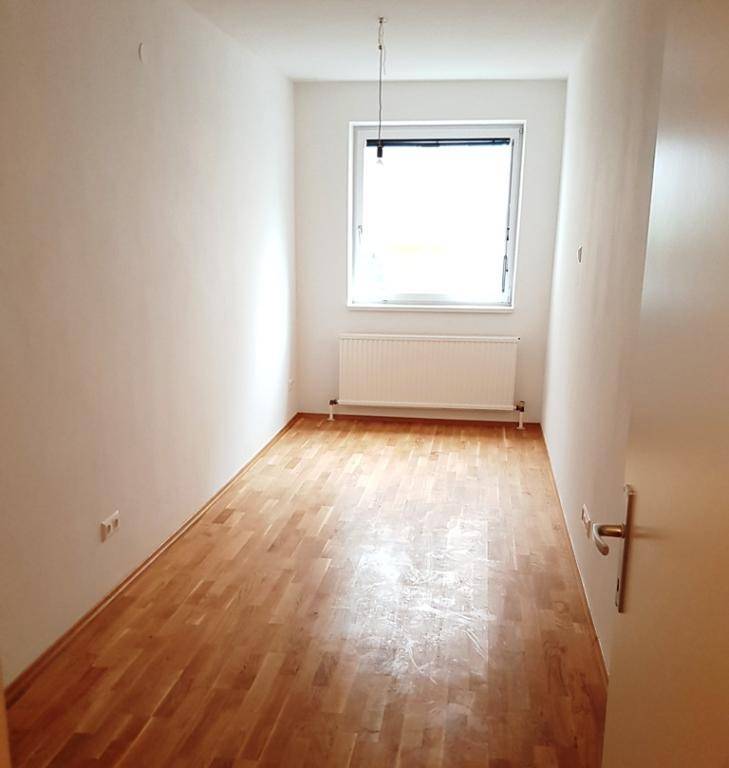 Helles, längliches Zimmer mit Parkettboden, weißen Wänden und einem Fenster mit Heizkörper.