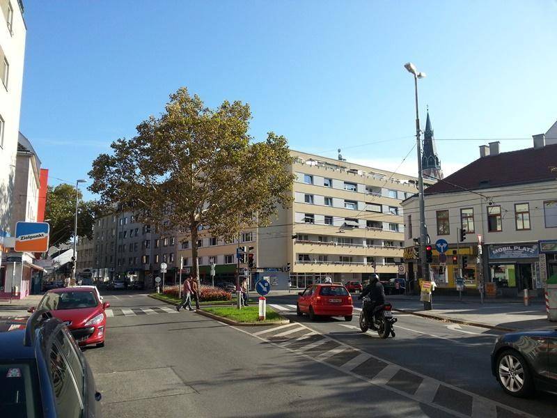 Belebte Stadtstraße mit Gebäuden, Bäumen und Fahrzeugen unter blauem Himmel, typisch urban.