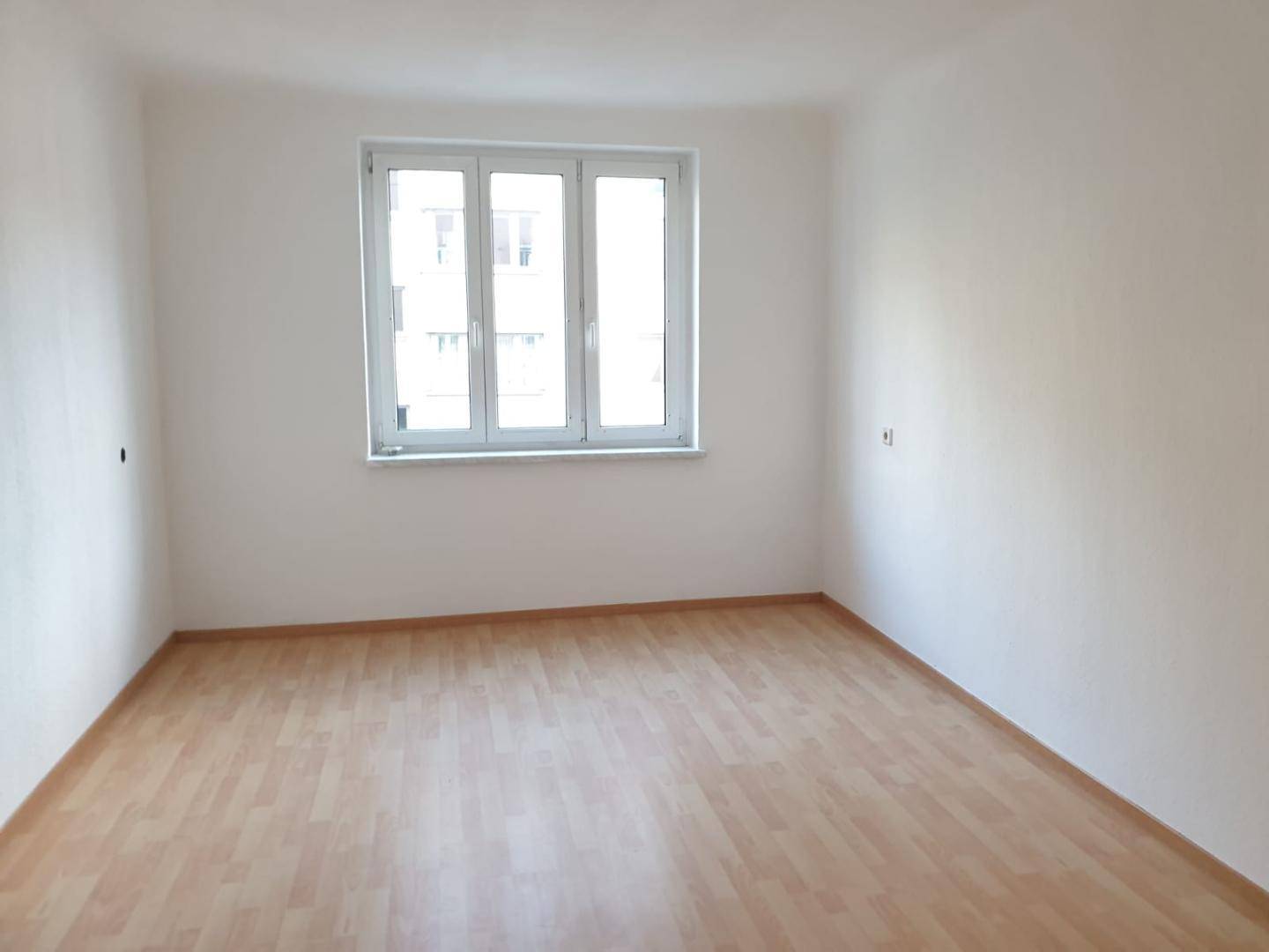 Leeres Schlafzimmer mit hellem Laminatboden und großem Fenster, das viel Tageslicht bietet.