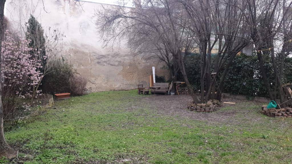 Hinterhofgarten mit Bäumen, Rasen und einer kleinen Sitzecke, umgeben von hohen Mauern.