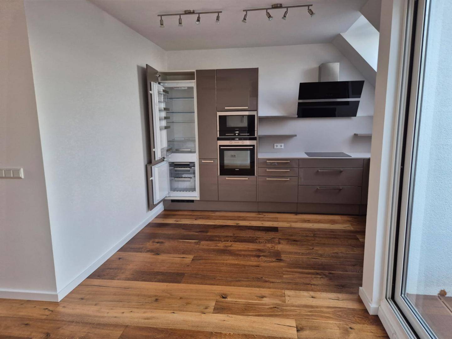 Moderne Einbauküche mit grauen Hochglanzfronten, integrierten Geräten und Holzboden in einem hellen Raum.