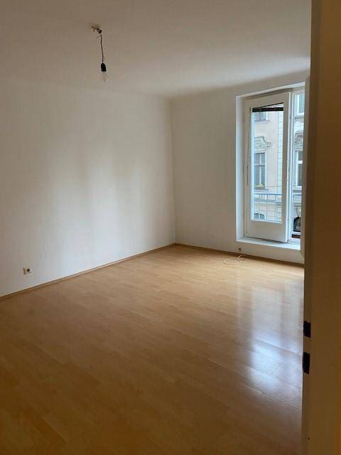 Geräumiges, leeres Zimmer mit Parkettboden und einem großen Fenster, das viel Licht spendet.