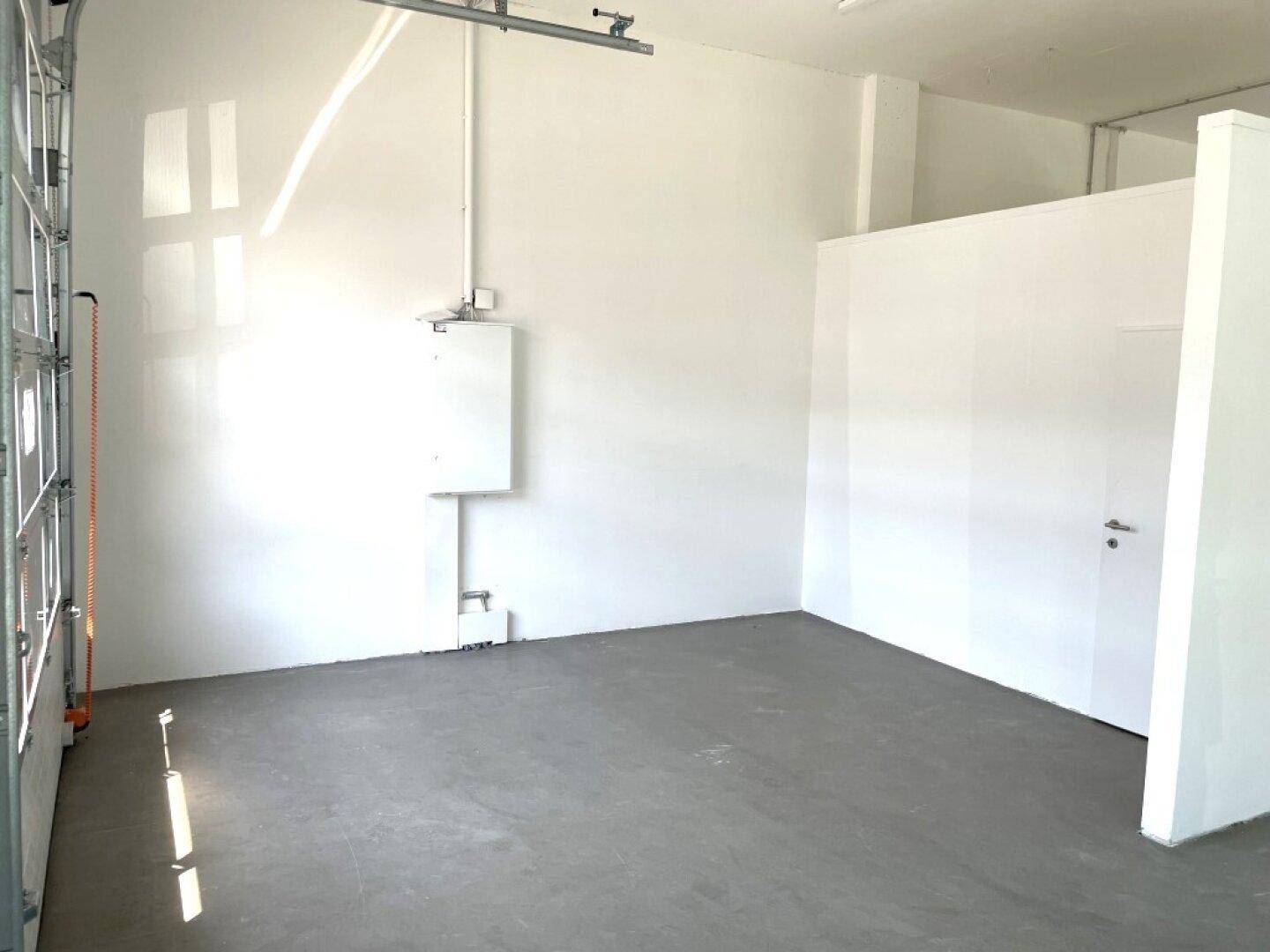 Geräumiger Gewerberaum mit Betonboden, weiß gestrichenen Wänden und einer Trennwand, die einen separaten Bereich schafft.
