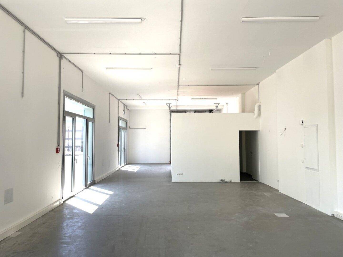 Weitläufiger, heller Gewerberaum mit Betonboden und sichtbaren Installationen an der Decke, bereit zur individuellen Gestaltung.