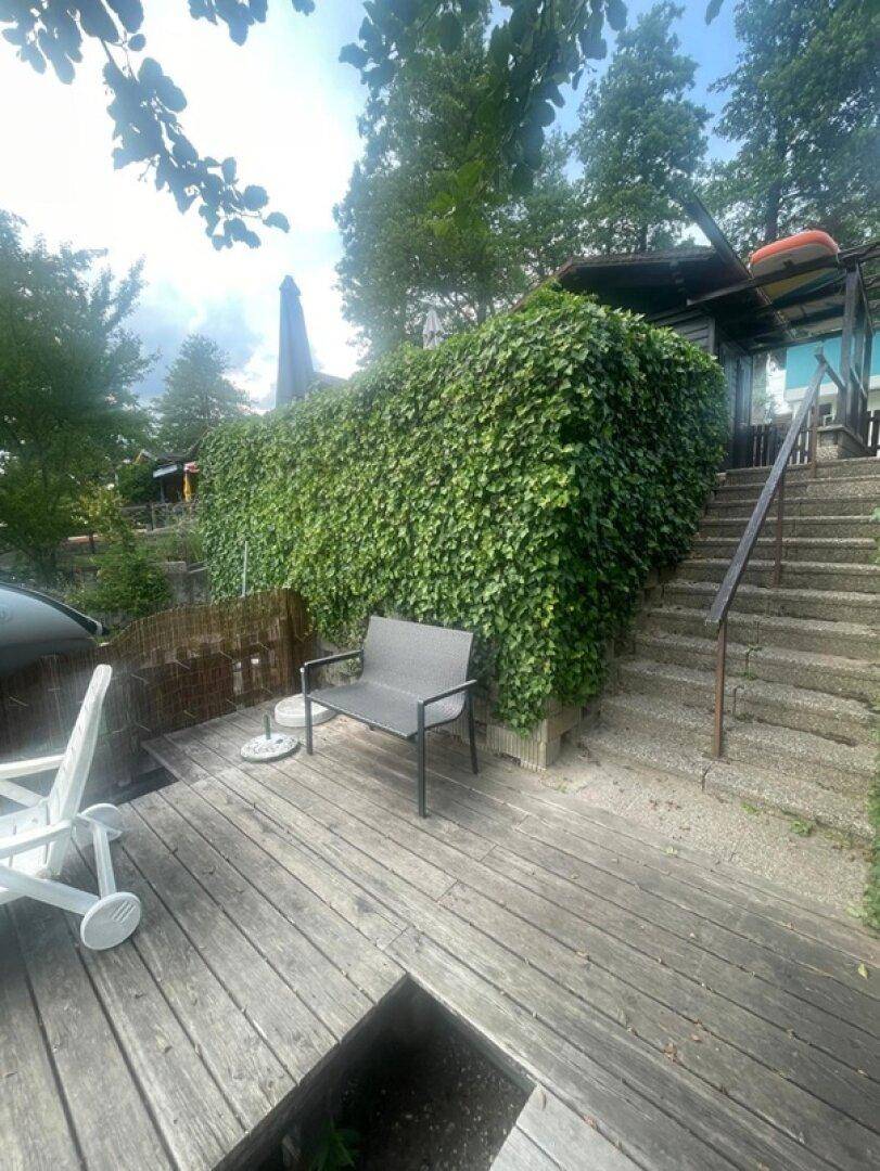 Holzterrasse mit Sitzbank und üppiger Begrünung, die zu einem Gebäude führt.