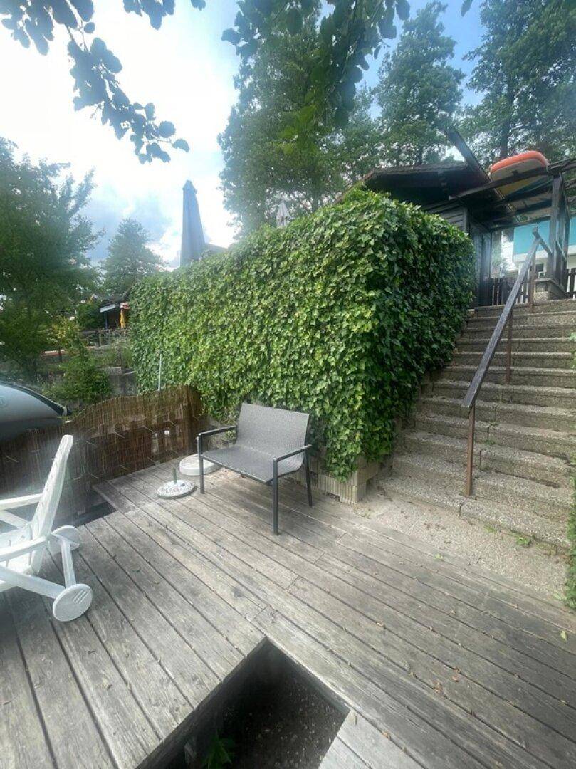 Holzterrasse mit Sitzbank und üppiger Begrünung, die zu einem Gebäude führt.