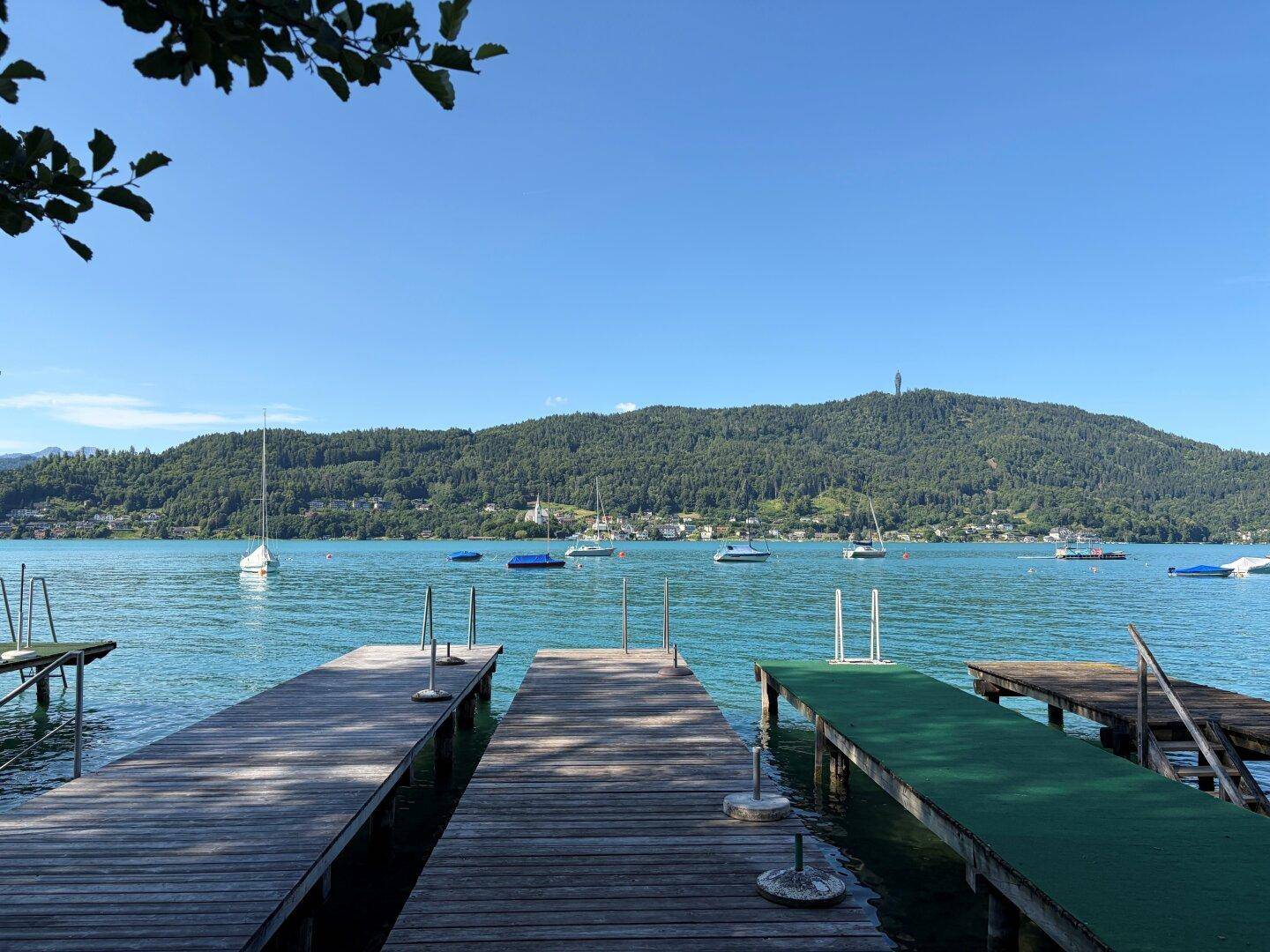 Weitläufiger Seeblick mit Holzstegen, Segelbooten und bewaldeten Bergen im Hintergrund.